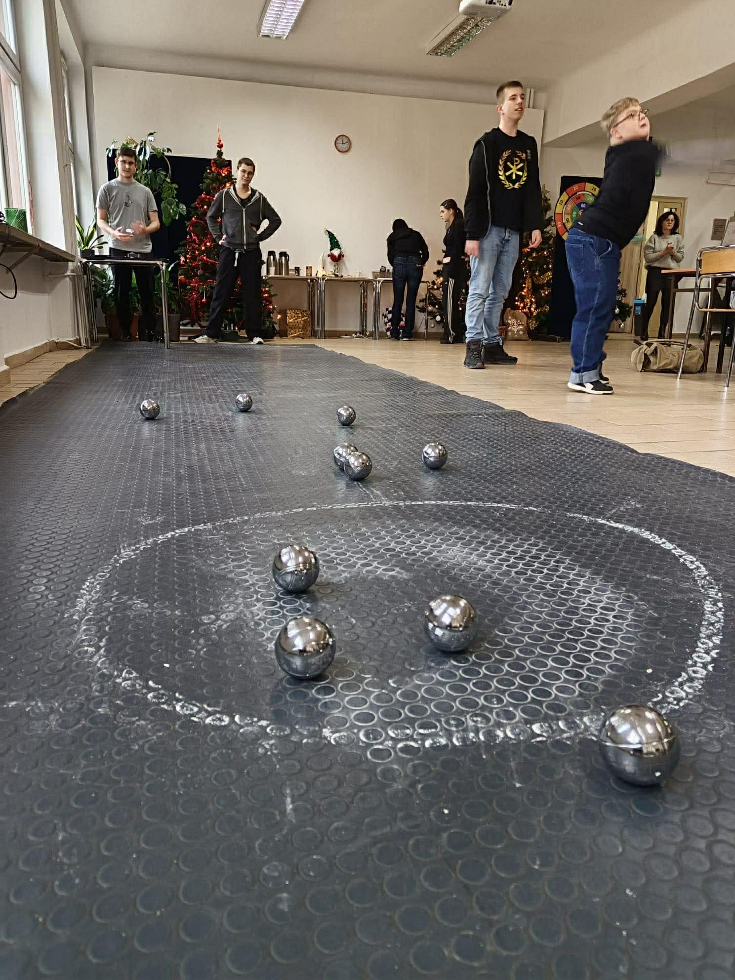 Metalowe kule do gry leżą na macie z zaznaczonym okręgiem. W tle młodzież gra w bule w sali udekorowanej choinkami i świątecznymi ozdobami.