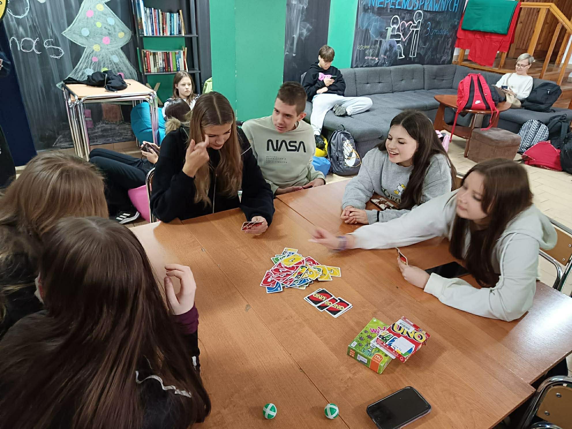 Grupa nastolatków siedzi przy stole i gra w karty UNO. W tle widać salę wspólną z kanapami, plecakami i tablicami z rysunkami.