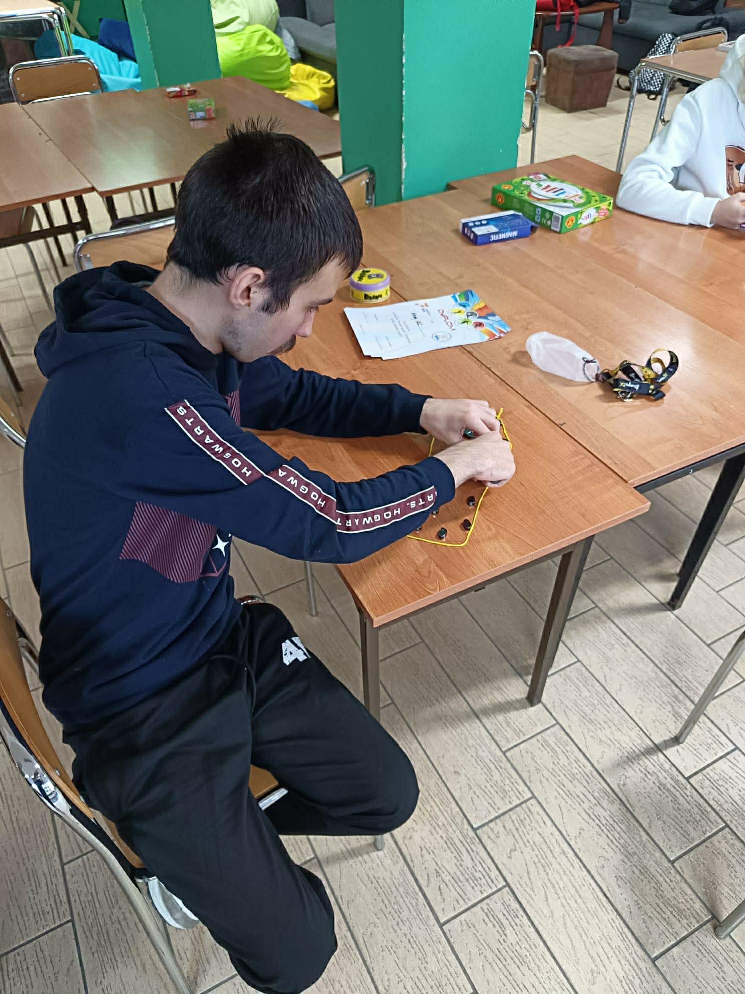 Młody mężczyzna siedzi przy stole i wykonuje proste prace manualne z użyciem drobnych elementów. Na blacie leżą materiały do zajęć, a w tle widać salę wspólną.